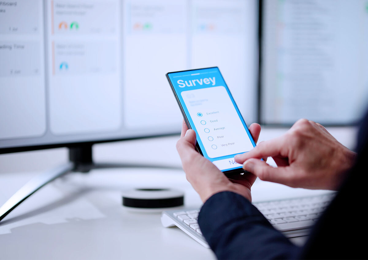 Close up of a person holding a mobile phone, which shows a survey on the screen. The background is blurred but you can make out computer monitors and a desk.