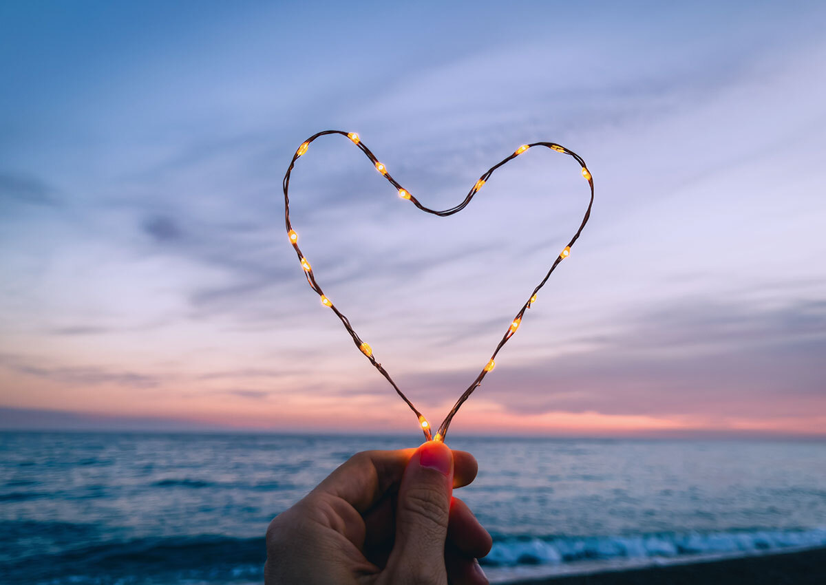 Branding: Values - photo of light string in the shape of a heart, being held by a person in front of an ocean view